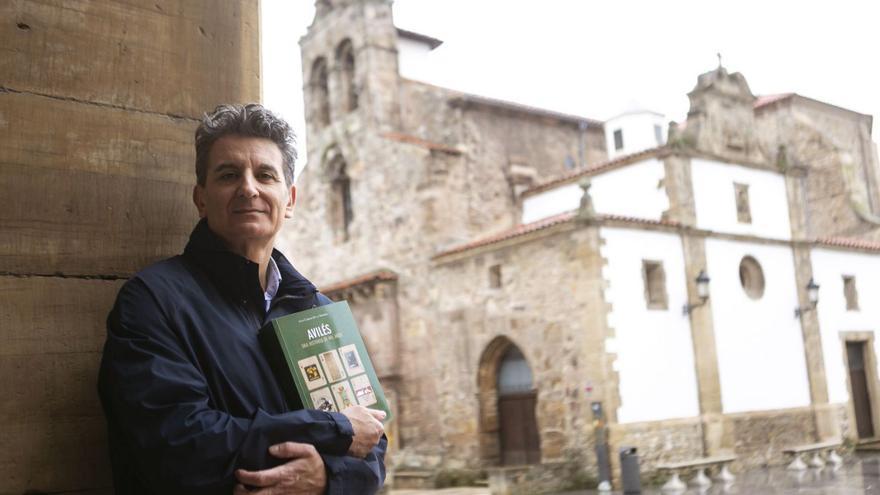 Juan Carlos de la Madrid posa en un soportal de la plaza Carlos Lobo con su último libro en la mano y la iglesia de San Antonio de Padua (el edificio más antiguo de Avilés) a su espalda. | María Fuentes