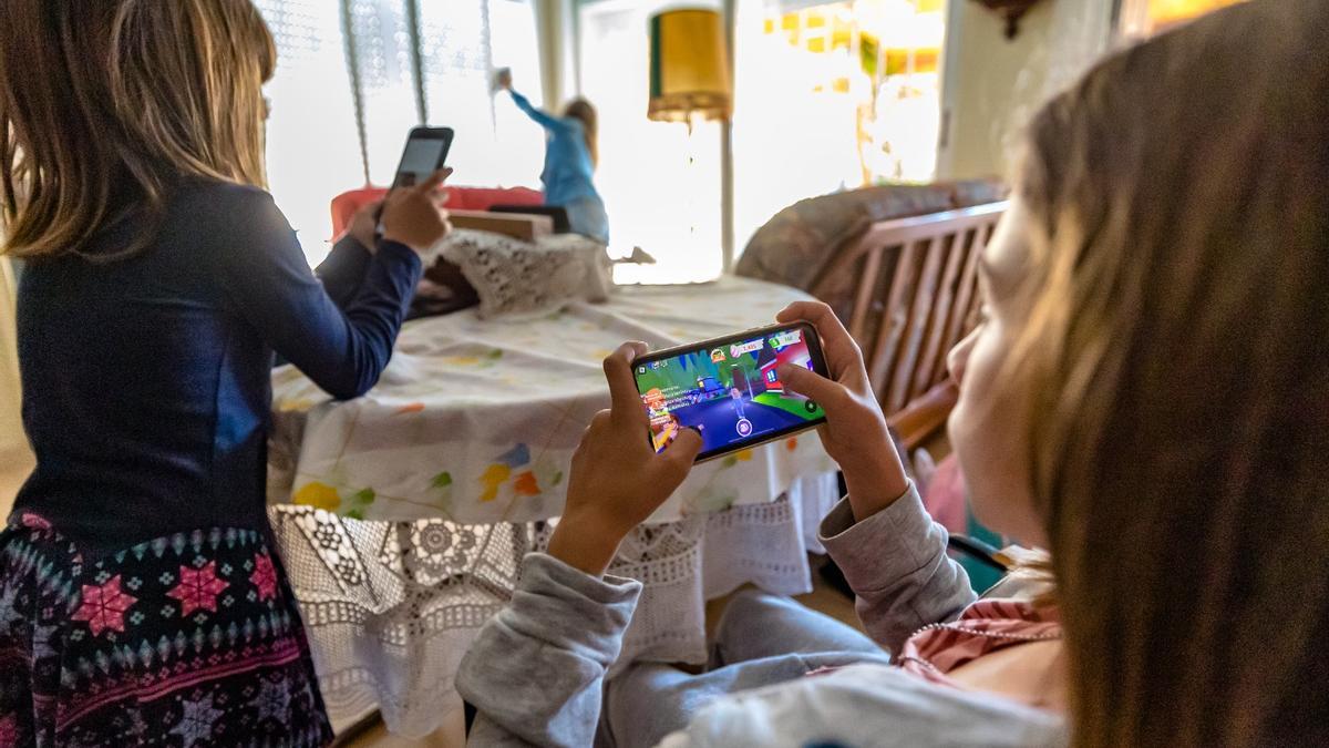 Dos niñas juegan con el teléfono de sus padres mientras estos realizan tareas domésticas.