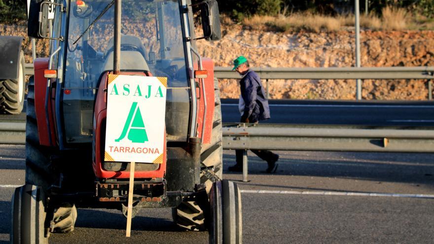 Un pagès passa per darrere d'un tractor amb un cartell d'ASAJA al tall de l'AP-7 a l'Ampolla