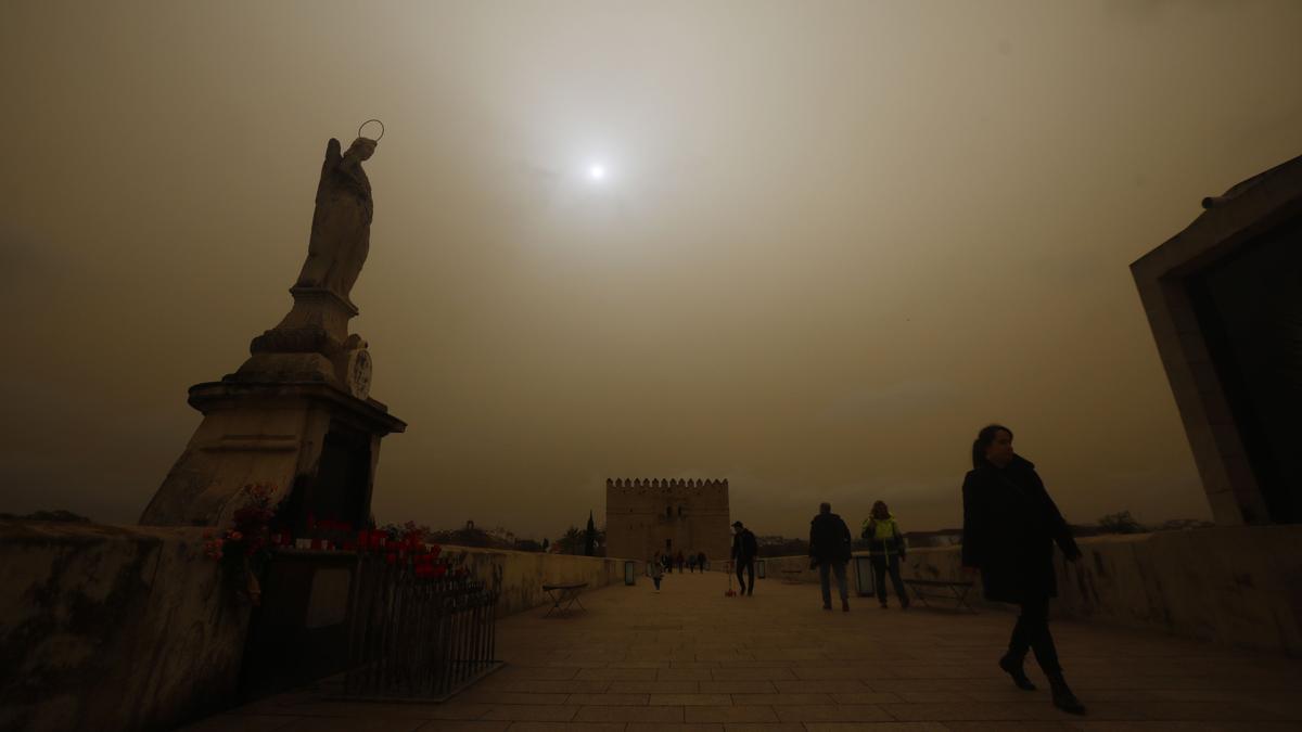 Varias personas caminan por el Puente Romano con la imagen del arcángel San Rafael de Córdoba con el cielo cubierto por la calima.