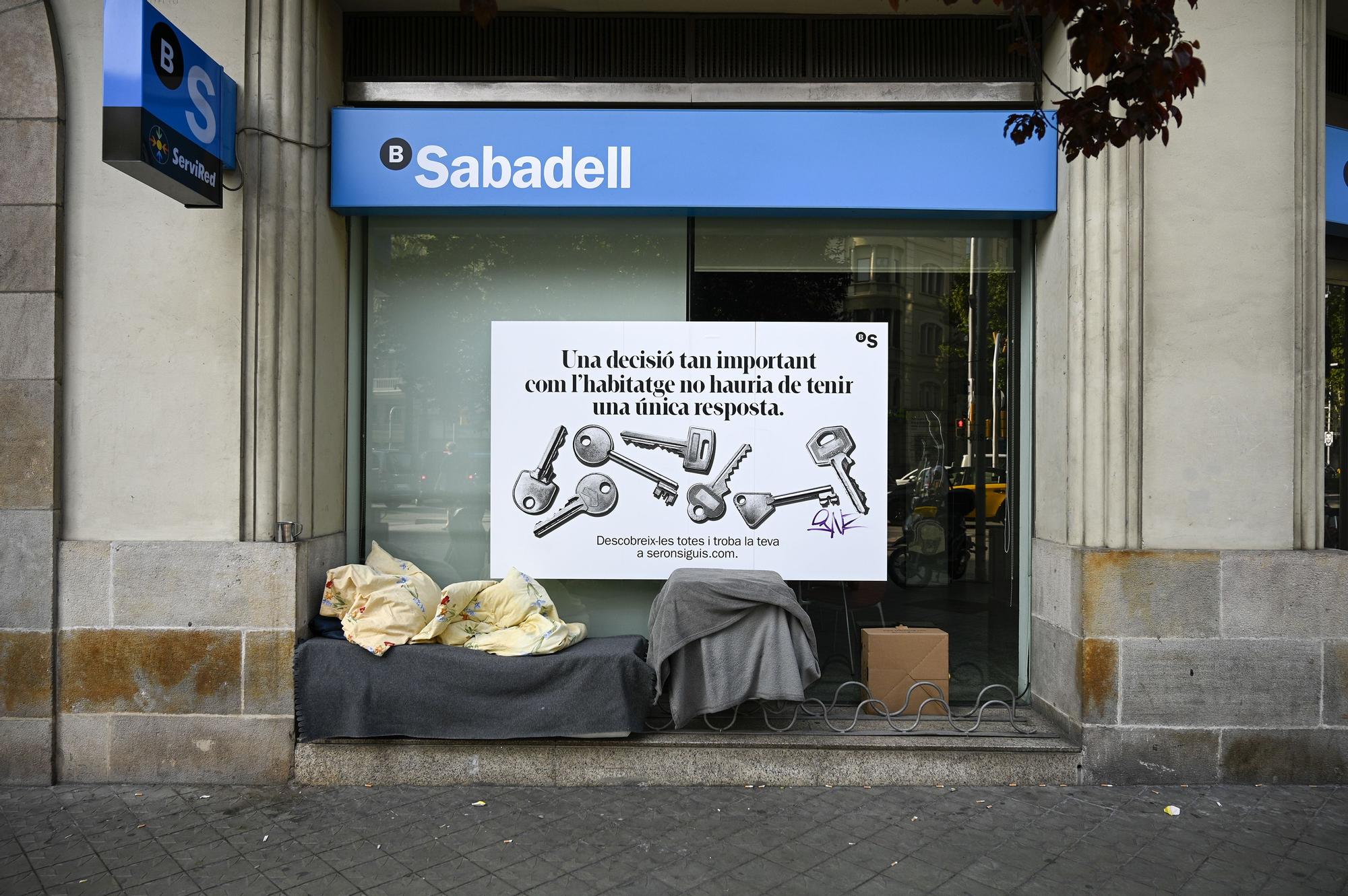 Enseres, probablemente de un 'sintecho', bajo la publicidad sobre la vivienda en un banco de Barcelona.