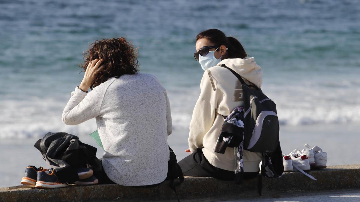 Dos mujeres con mascarilla charlando en Samil frente al mar.