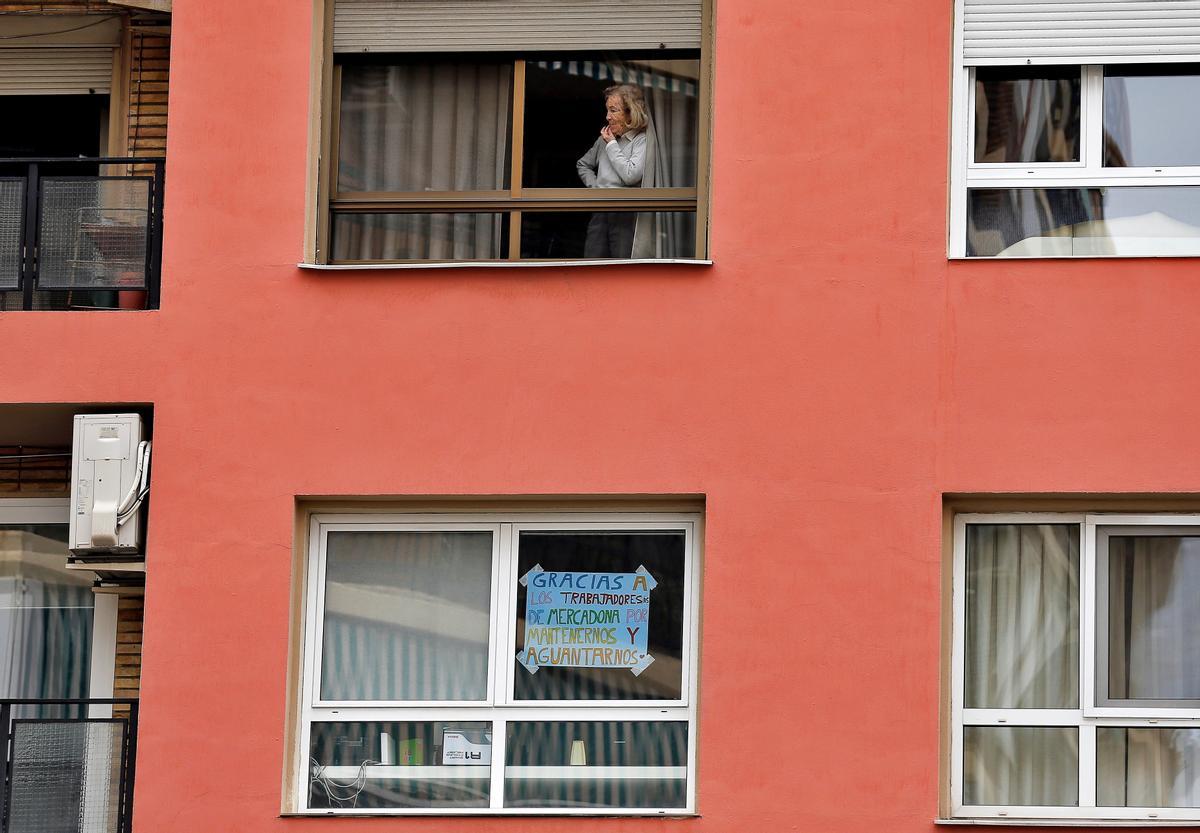 360. VALENCIA, 23/04/2020.- Una mujer observa la calle desde la ventana de su casa, mientras otro vecino ha colocado un cartel de apoyo a todos los trabajadores de un superermercado cercano, este jueves que se cumplen los cuarenta días de confinamiento debido a la pandemia provocada por el