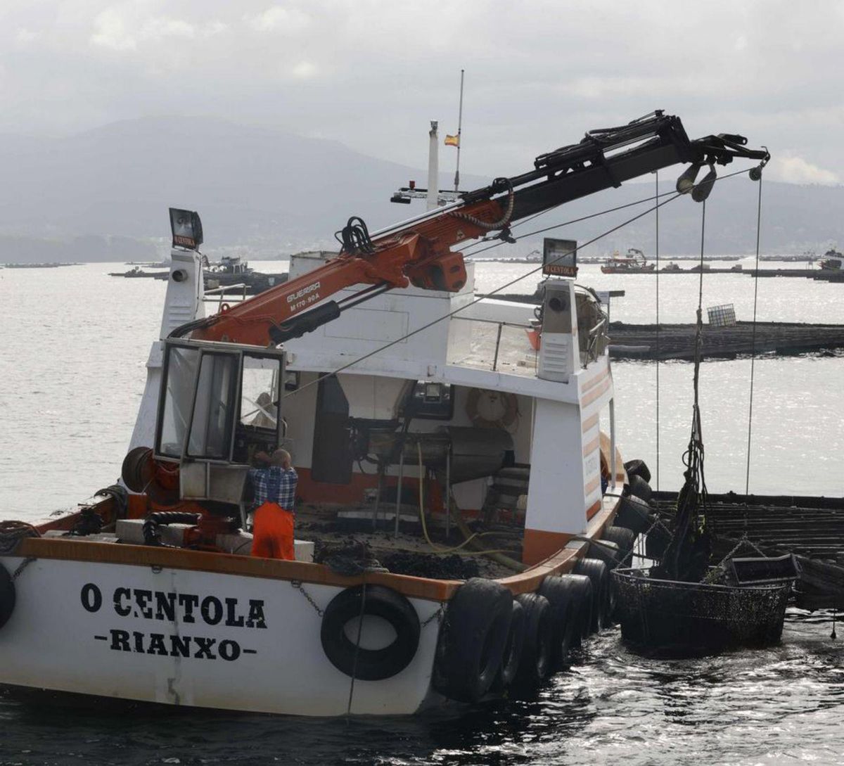 Un barco auxiliar de acuicultura se ayuda de la grúa y el cesto para retirar cuerdas de una batea. | // PABLO HERNÁNDEZ