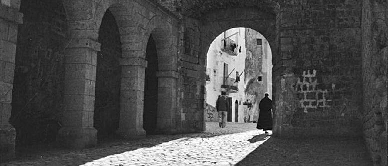 Dos varones cruzan bajo el arco de la plaza de Vila, uno de ellos vestido con sotana.