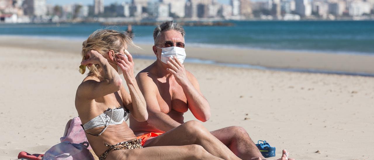 Dos personas tomando el sol con mascarilla en la playa del Postiguet de Alicante.