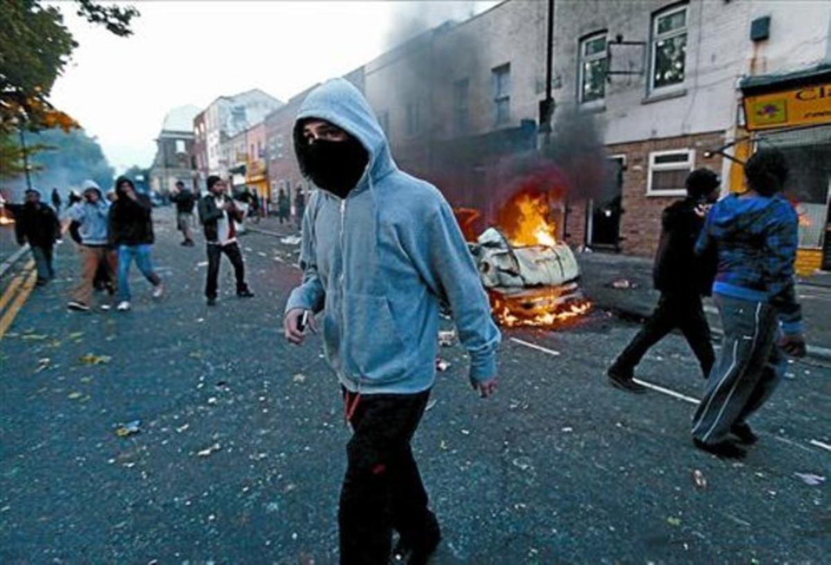 Vehicles incendiats 8 Un noi amb el seu mòbil davant d’un cotxe en flames al barri de Hackney. Vehicles incendiats 8 Un noi amb el seu mòbil davant d’un cotxe en flames al barri de Hackney.