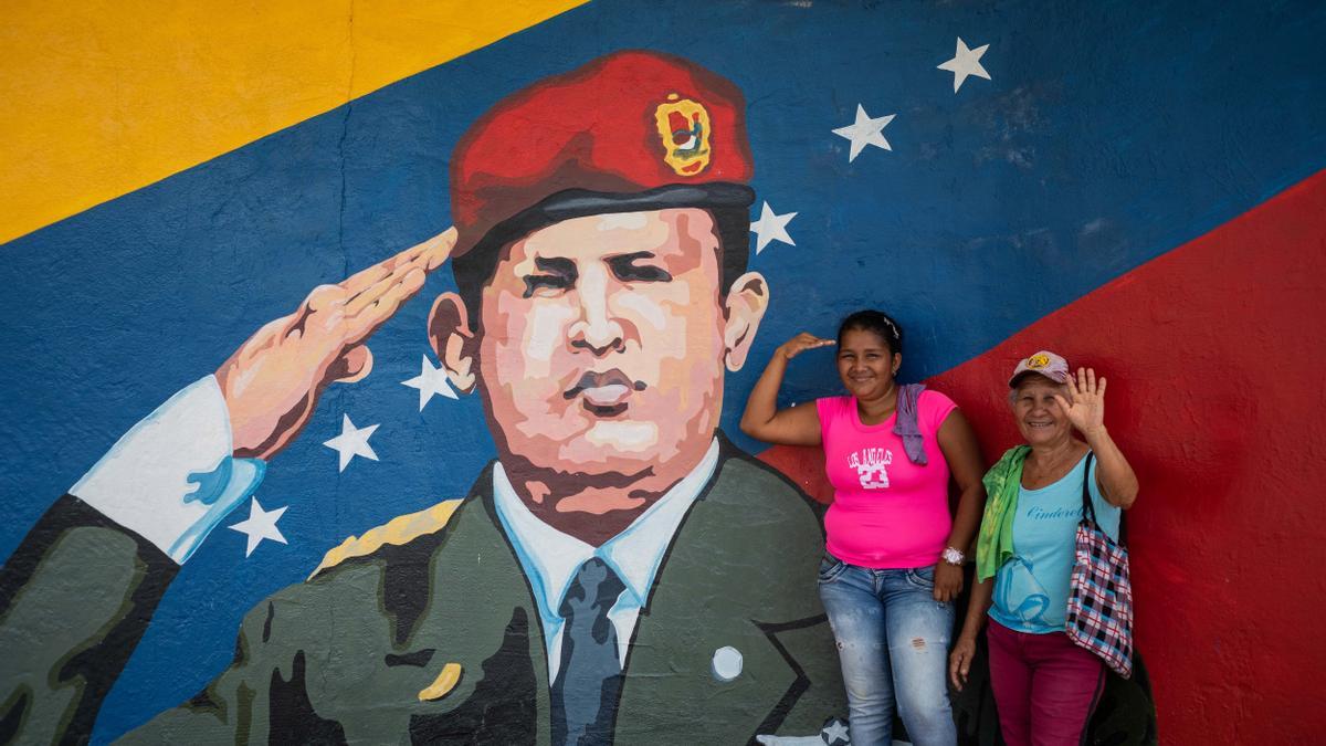 Dos mujeres posan para una fotogarfía junto a un mural de Hugo Chávez