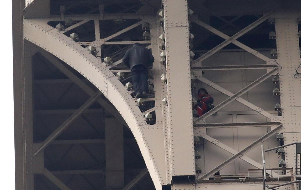 El escalador sube por la estructura de la Torre Eiffel con un bombero vigilante a pocos metros.