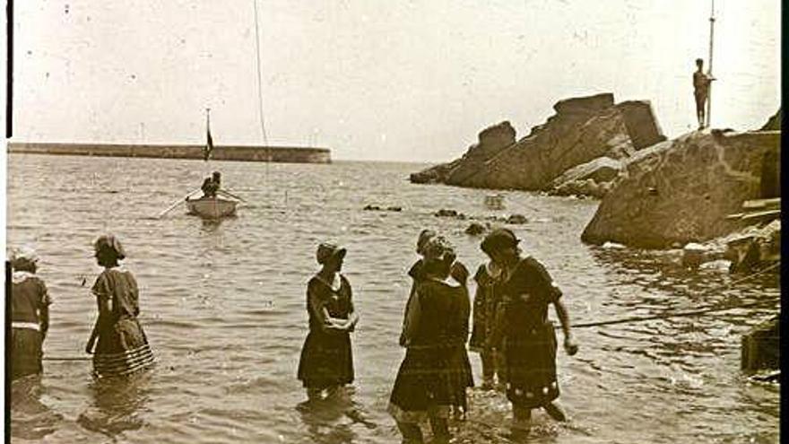 Gentada a la terrassa dels banys de Sant Elm en una regata de patí català. Dones protegint-se del sol, banyant-se a la platgeta dels banys de Sant Elm, entre 1922-1929.