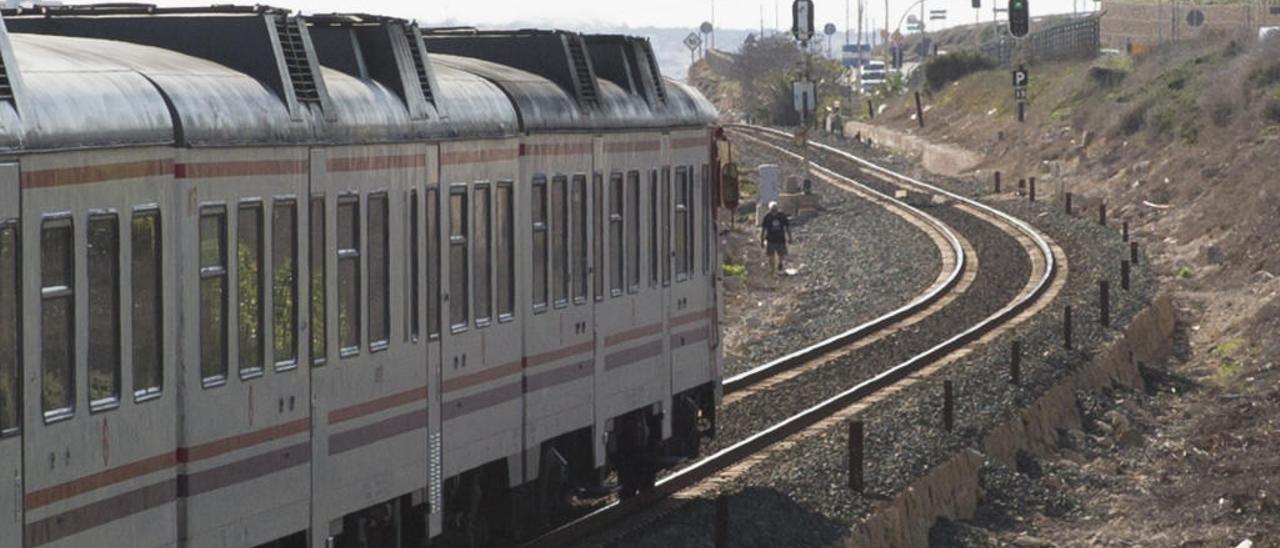 Una persona cruza las vías frente a la valla de seguridad agujereada mientras circula un tren de cercanías.