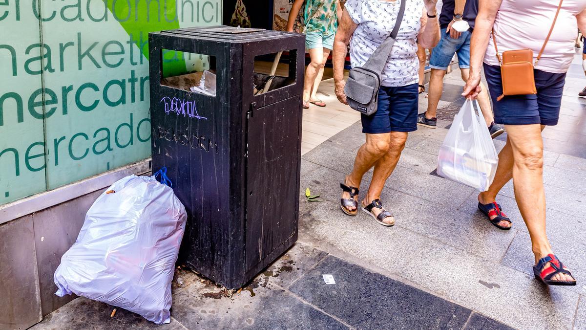 Una bolsa de basura junto a una papelera hasta arriba de desperdicios y llena de mugre, junto al mercado municipal de Benidorm y a pleno día.