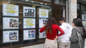 Jóvenes miran ofertas de alquiler en el escaparate de una oficina inmobiliaria, en Santiago de Compostela. / EFE/ Xoán Rey