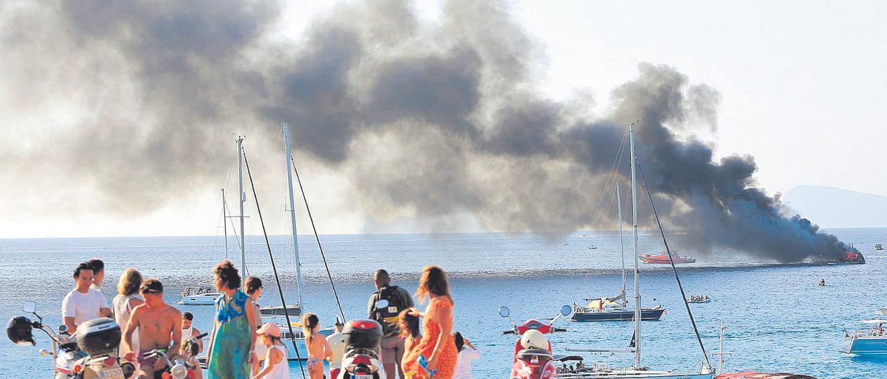 Bañistas en la costa de Cala Saona y, al fondo, el yate envuelto en llamas.