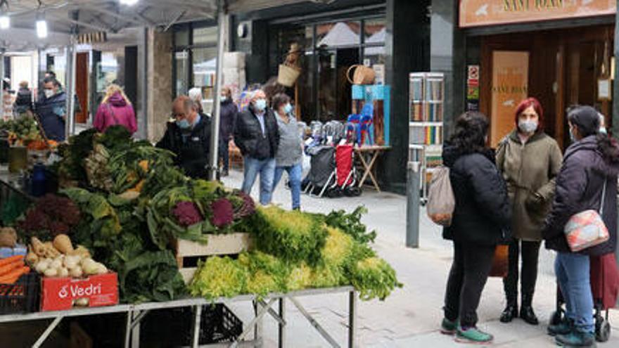 Parada del mercat setmanal de la Seu d&#039;Urgell, clients adquirint fruites i verdures i, al fons, alguns dels comerços del carrer Major el 2 de març de 2021