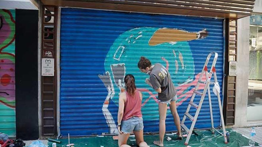 La artista JÃºlia Mulet y una ayudante pintan la barrera de una de las tiendas de las galerÃ­as Geranis.