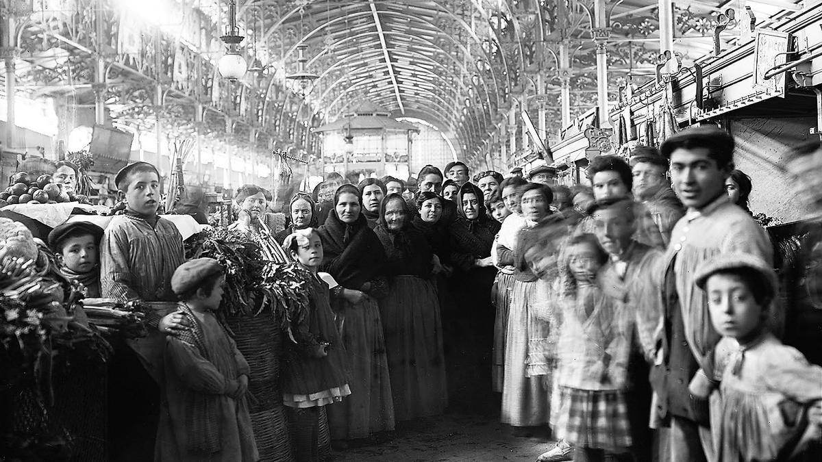 Interior del Nuevo Mercado construido sobre una parte de la antigua plaza del Mercado, en una imagen que se centra sobre un antropológico grupo de vendedoras de verduras y otros productos de la huerta de proximidad, flanqueadas por mozos y vivaces pardales en 1912