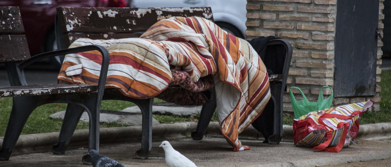Una persona sin hogar duerme en un banco de una calle de Alicante