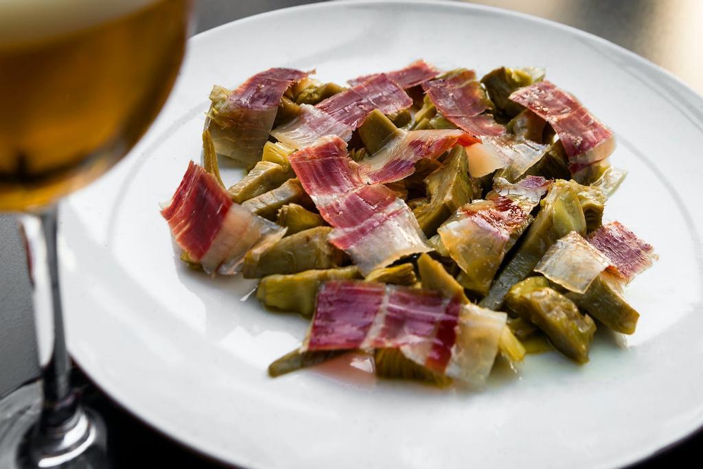 Alcachofas naturales con jamón ibérico: primero, pelar las alcachofas, dejar solo el corazón y cortar en cuatro trozos; freír en aceite de oliva virgen y reservar. En el momento de servir, hornear y decorar con jamón ibérico de bellota.