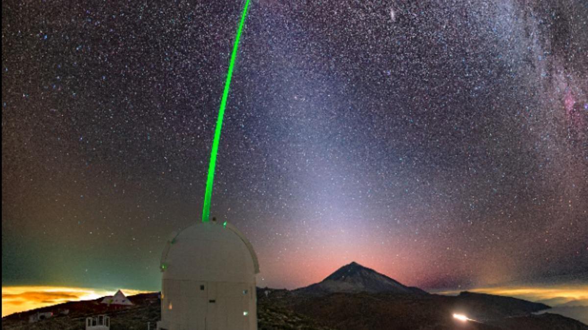 Estación óptica terrestre propiedad de la Agencia Espacial Europea (ESA), ubicada en el Observatorio del Teide del Instituto de Astrofísica de Canarias. Con el Teide al fondo y con un cielo plagado de las estrellas de nuestra Galaxia, la Vía Láctea, se lanza un haz láser de un metro diámetro utilizando completamente la apertura del telescopio. Fotografía realizada durante los experimentos de corrección de óptica adaptativa inversa, uplink, durante el doctorado de Noelia Martínez, dirigida por Luis Fernando Rodríguez Ramos, donde ese tipo de corrección se consiguió por primera vez a nivel mundial. El haz láser aparece curvado por efecto del objetivo gran angular con el que se tomó la fotografía.
