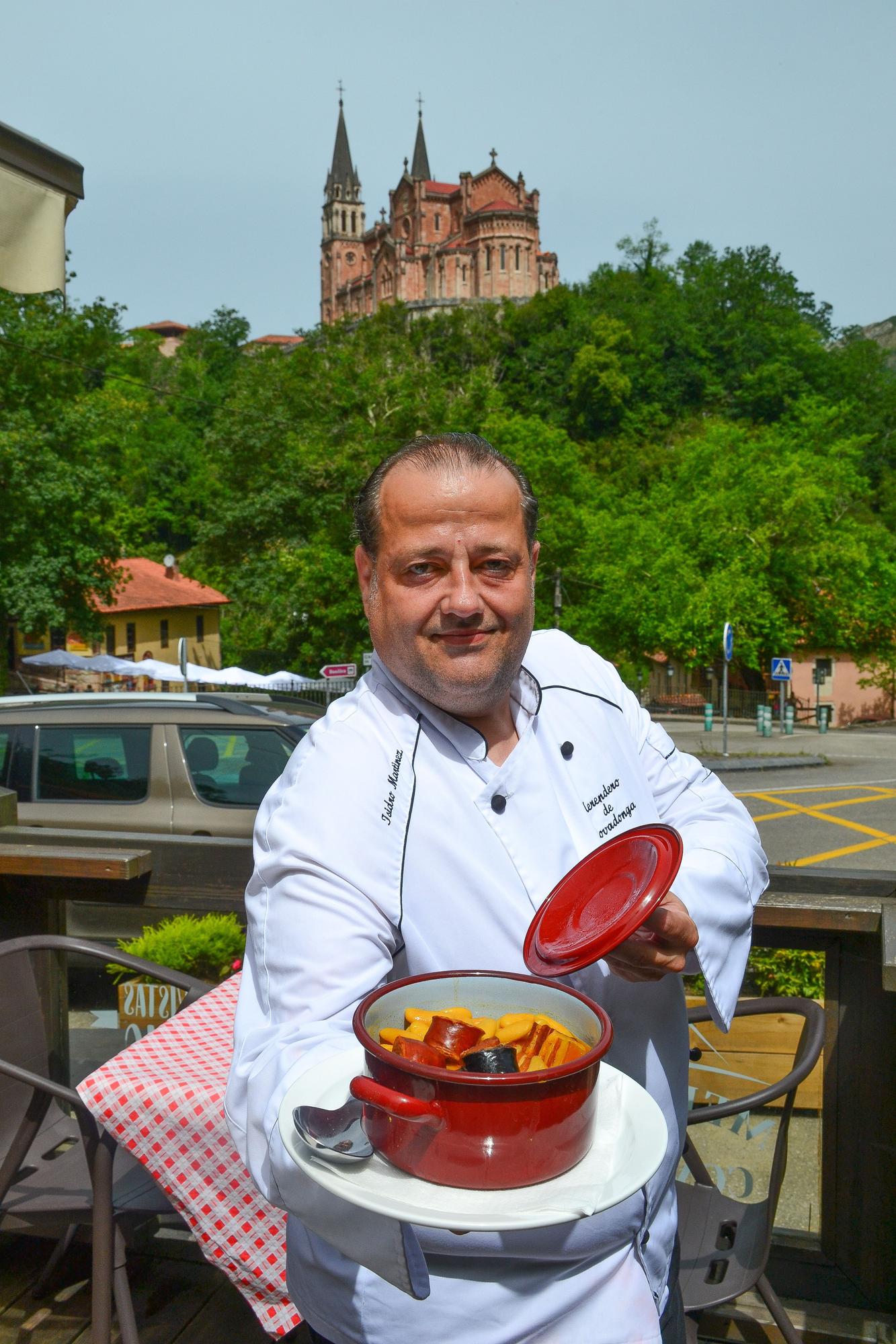 Isidro Martínez muestra la fabada que cocina en su restaurante, con el santuario de Covadonga a sus espaldas.