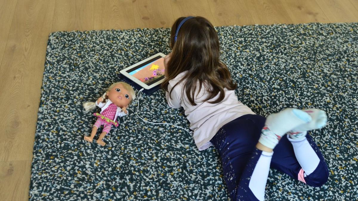 Una niña se entretiene en casa con una tablet y muñecas.
