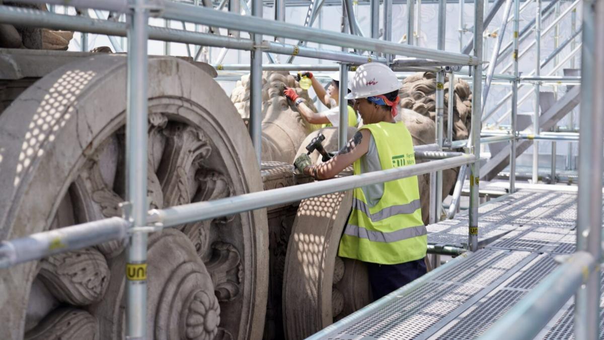 Dos restauradoras trabajando entre los andamios junto a las ruedas del carro y los leones de la Fuente de Cibeles.
