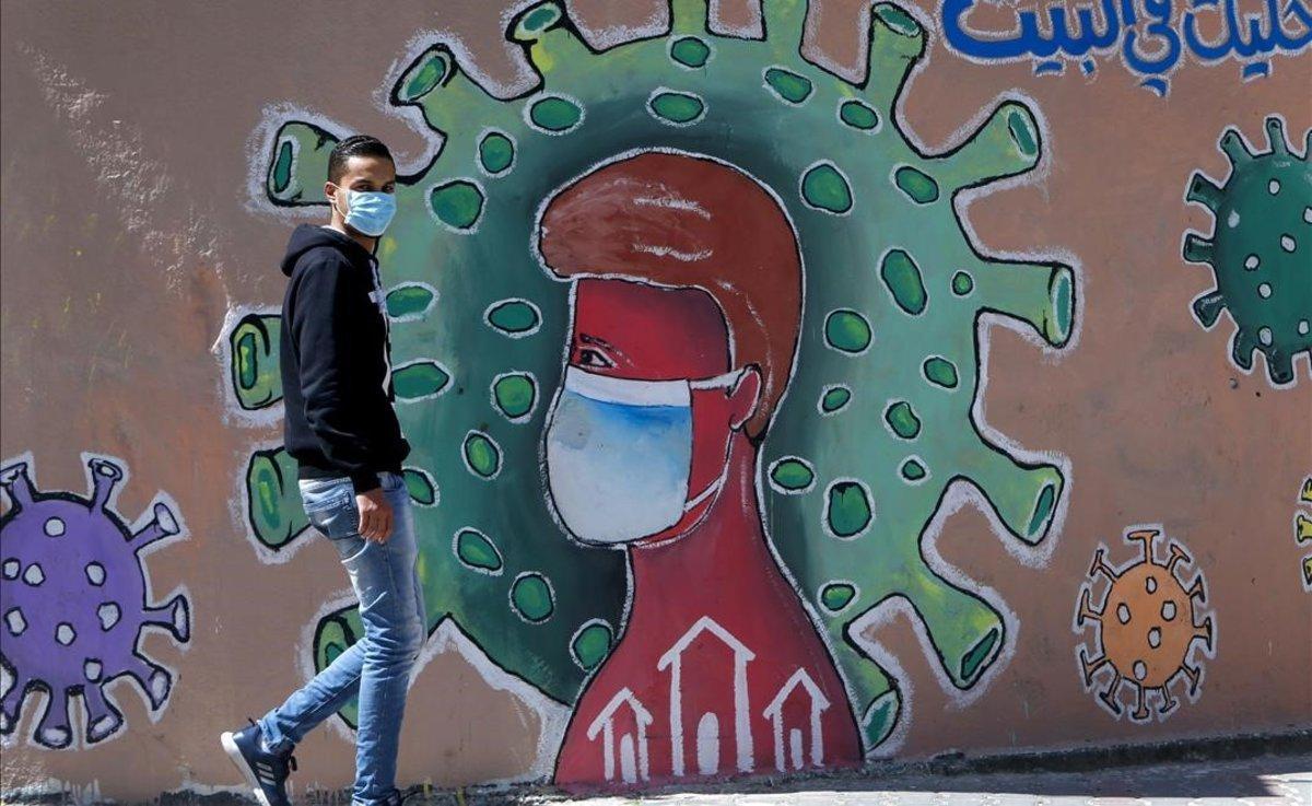 Un hombre con mascarilla pasa frente a un mural alusivo a la pandemia de coronavius, en Gaza.