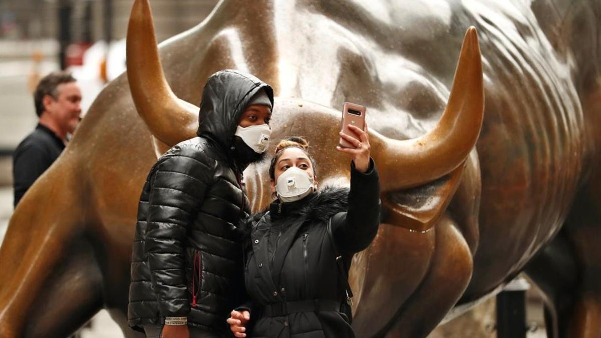Dos jóvenes con mascarilla se hacen un 'selfie' frente a la estatua del toro de Wall Street, este martes en Nueva York.