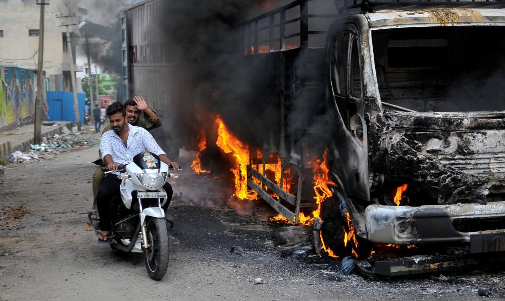 Hombres montan en una motocicleta al lado de un camión en Bangalore, que fue incendiado por los manifestantes en la India.