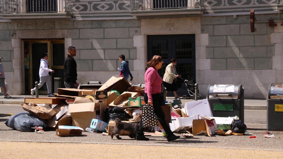 Persones caminant a Figueres al costat d'uns contenidors de deixalles desbordats.