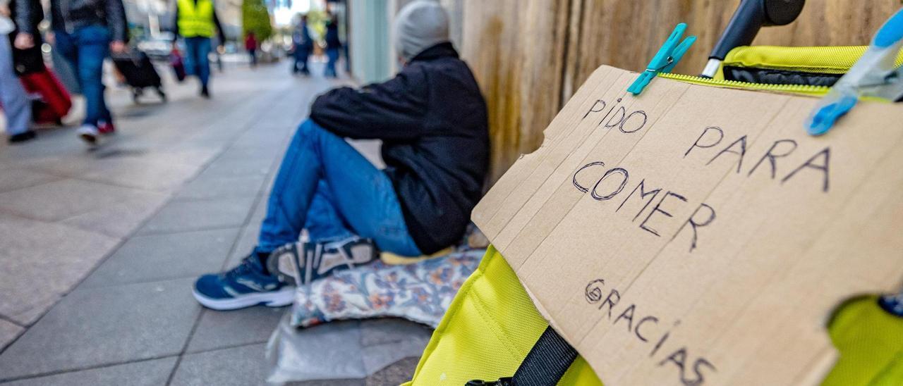 Un hombre, sentado sobre una acera, pide limosna a los viandantes.