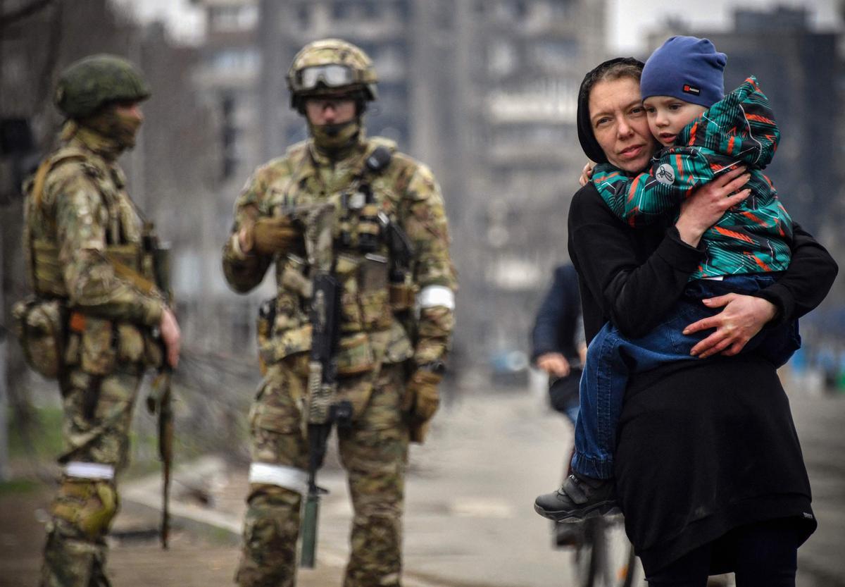 12 de abril de 2022.- Una mujer pasa con un niño junto a dos soldados rusos que ocupan la ciudad de Mariúpol, en el este de Ucrania.