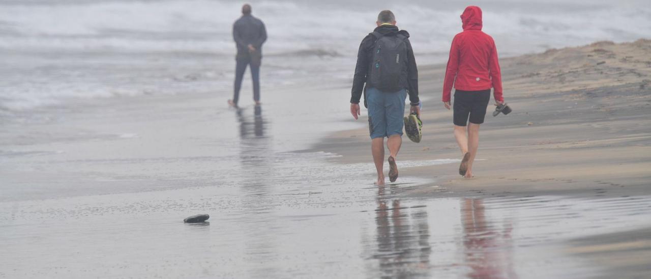 Dos turistas pasean por una playa mientras un tercero otea el horizonte.