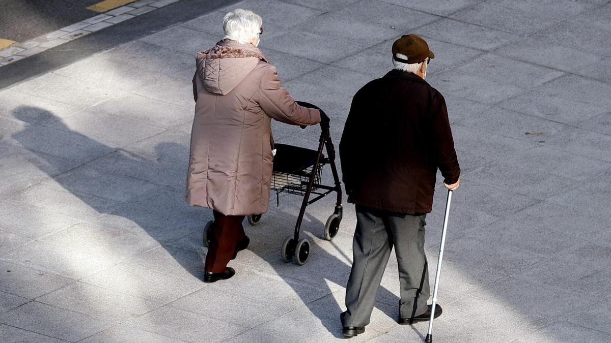 Dos personas mayores caminan con apoyo de un andador y un bastón.