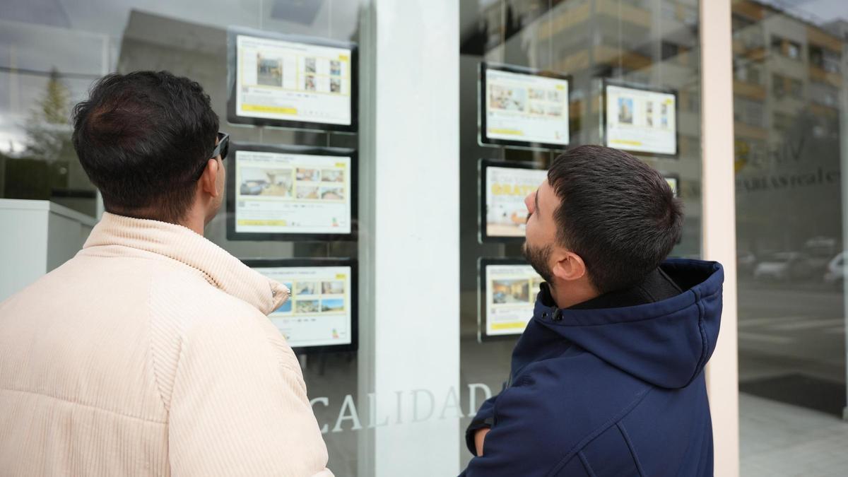 Dos jóvenes observan anuncios de pisos en venta en el escaparate de una inmobiliaria en Badajoz.