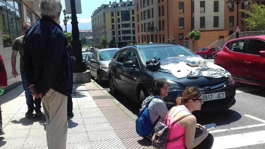 Vecinos sentados en la acera junto al coche en el que quedó atrapado el felino, con el capó cubierto con objetos blancos.