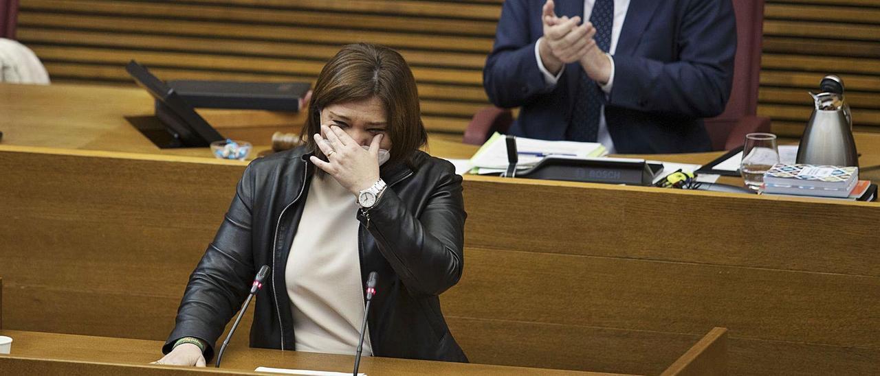 Isabel Bonig, ayer, emocionada durante su discurso de despedida en las Cortes Valencianas, con Morera al fondo aplaudiendo. | FERNANDO BUSTAMANTE