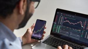 Business man trader investor analyst using mobile phone app analytics for cryptocurrency financial market analysis, trading data index chart graph on smartphone and laptop screen. Over shoulder view