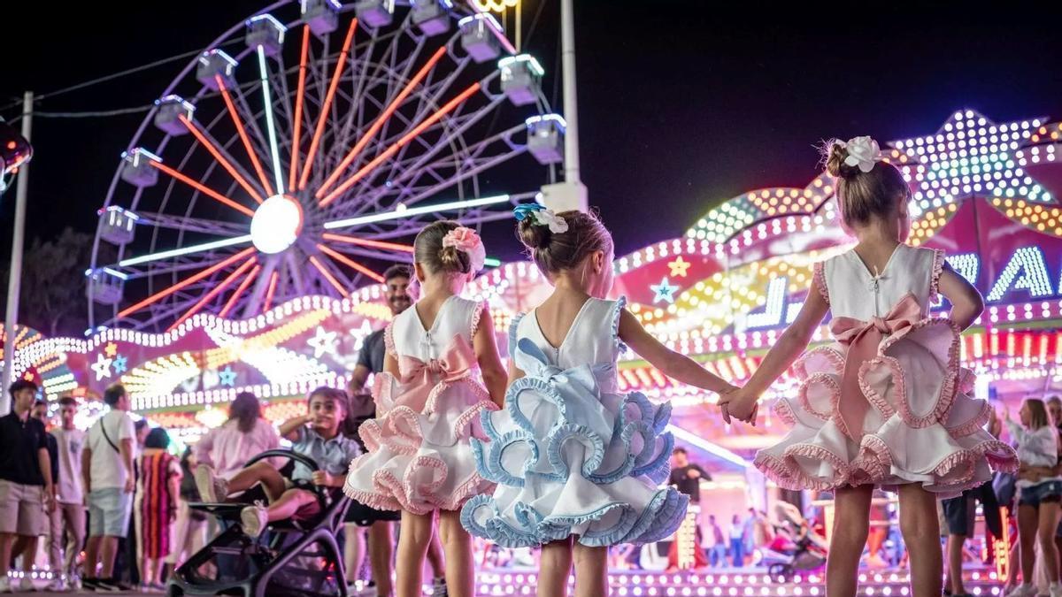 Tres niñas contemplan las atracciones del ferial con la noria al fondo, una de las imágenes más reconocibles de años anteriores.