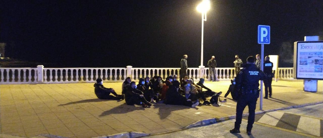 Inmigrantes que han llegado en patera a la playa de Los Locos de Torrevieja, sentados en el paseo marítimo y custodiados por la Policía Local