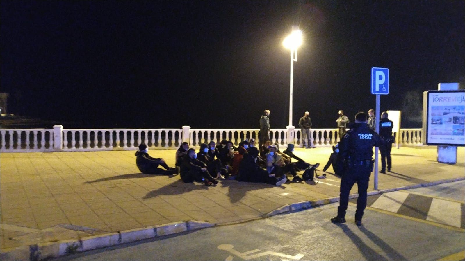 Inmigrantes que han llegado en patera a la playa de Los Locos de Torrevieja, sentados en el paseo marítimo y custodiados por la Policía Local