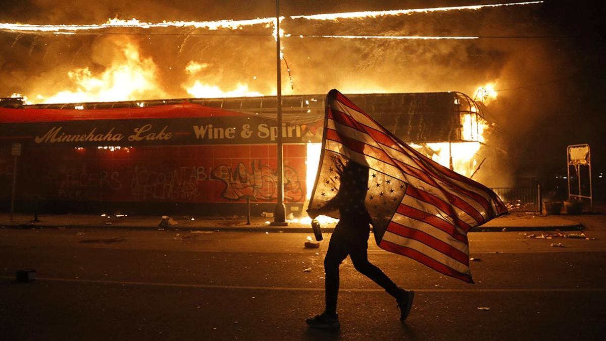 Un manifestante porta una bandera estadounidense al revés, junto a un edificio en llamas, en Minneápolis el jueves 28 de mayo