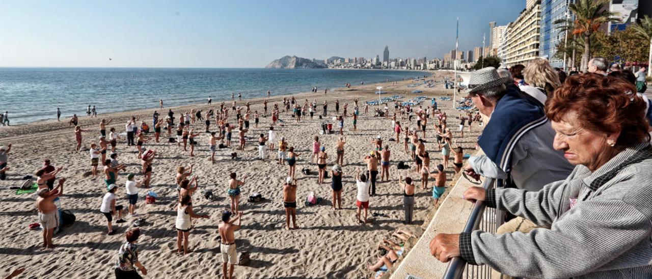 Turistas mayores observan a otros jubilados haciendo gimnasia en la playa de Poniente de Benidorm el pasado invierno.