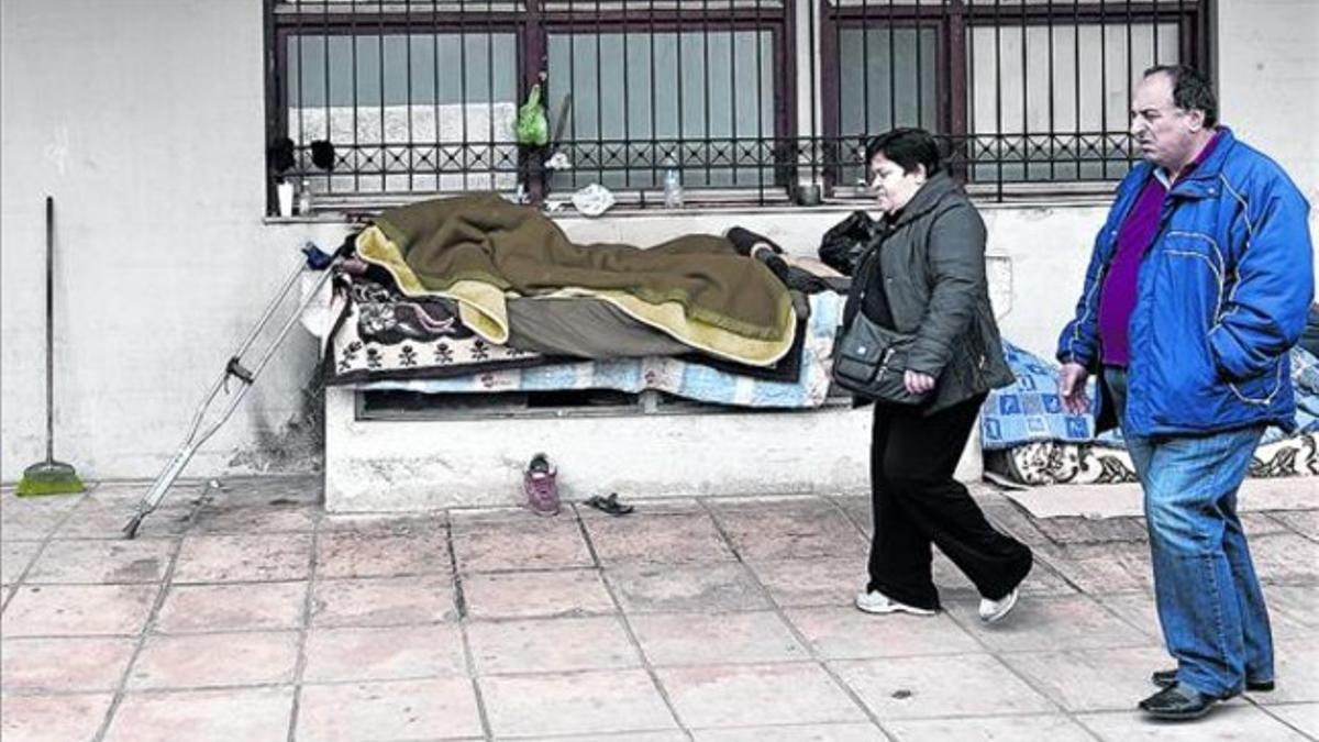 Dos ciudadanos pasan junto a un 'sin techo' durmiendo en un banco, en Salónica, este jueves.