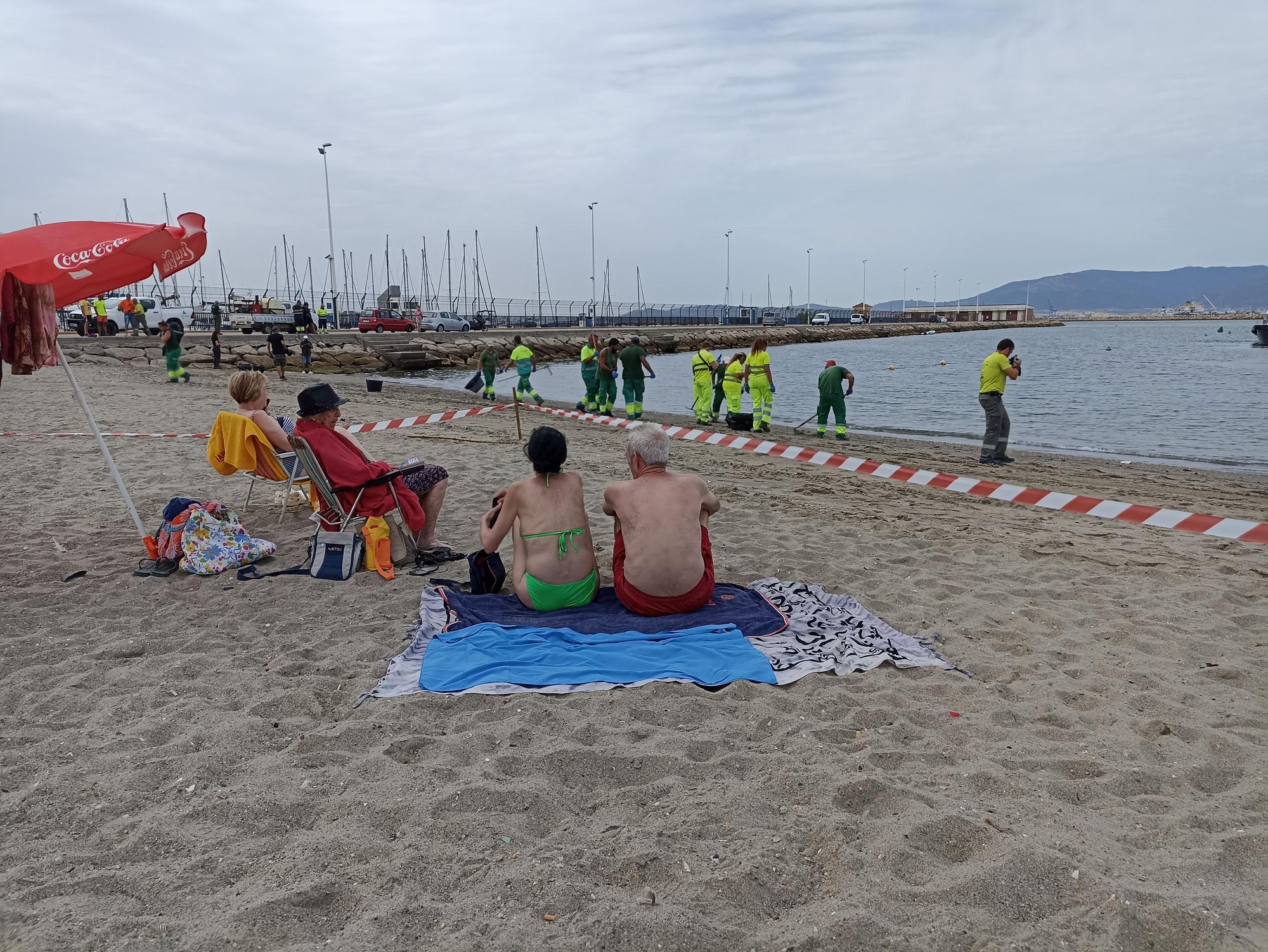 Turistas observan como operarios de La Línea retiran restos de aceite de la Playa de Poniente.
