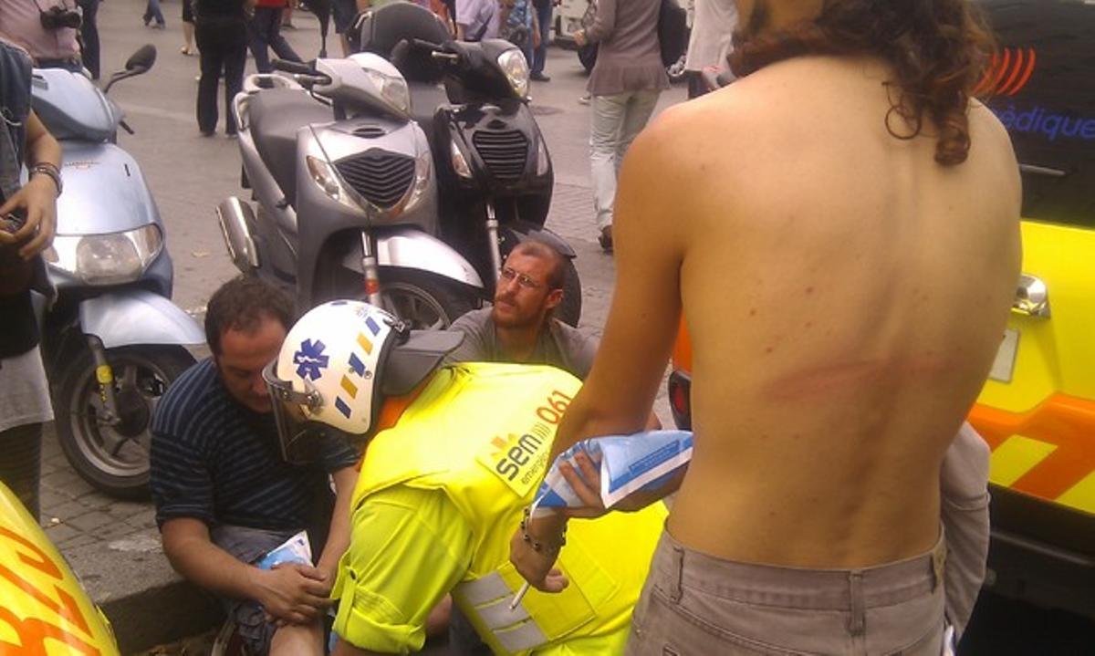 Roc Peris mostra el senyal del cop de porra a l’esquena mentre personal sanitari atén David Fernández, que ha patit contusions a les cames.