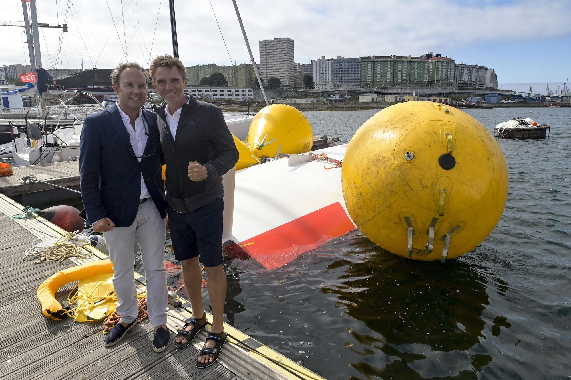 El navegante francés Laurent Camprubi (d), acompañado por el Jefe de Salvamento Marítimo Finisterre, Manuel Capeáns (i), posan delante del velero volcado y amarrado en el pantalán del puerto marítimo de Oza