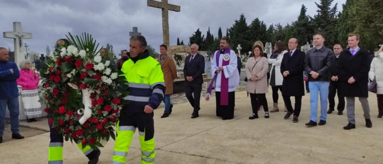 Dos operarios municipales llevan una corona de flores a la cruz del paseo central.