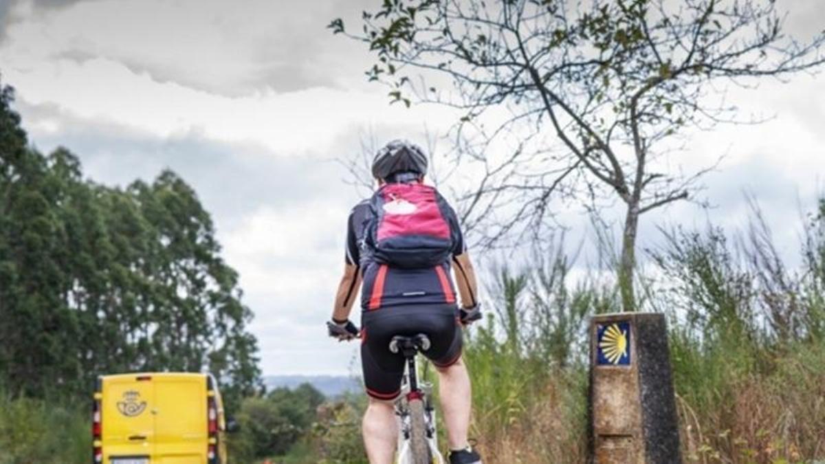 Un peregrino recorre en bicicleta una etapa del Camino de Santiago junto a una señal jacobea y un vehículo de Correos.